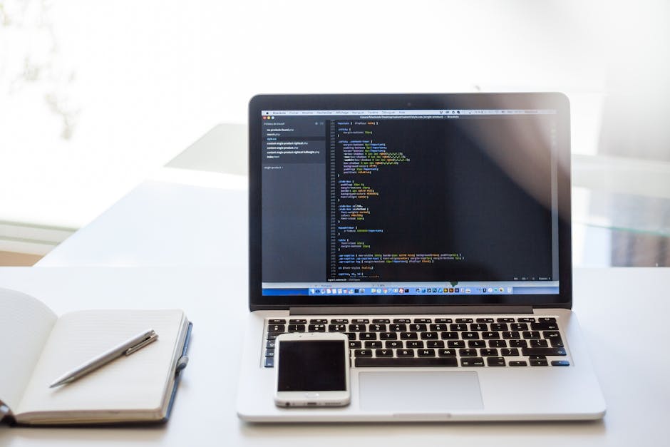 A laptop computer displaying lines of code on its dark screen, placed on a white desk with a closed notebook and a pen to the left, and a smartphone positioned in front of the laptop. The workspace is